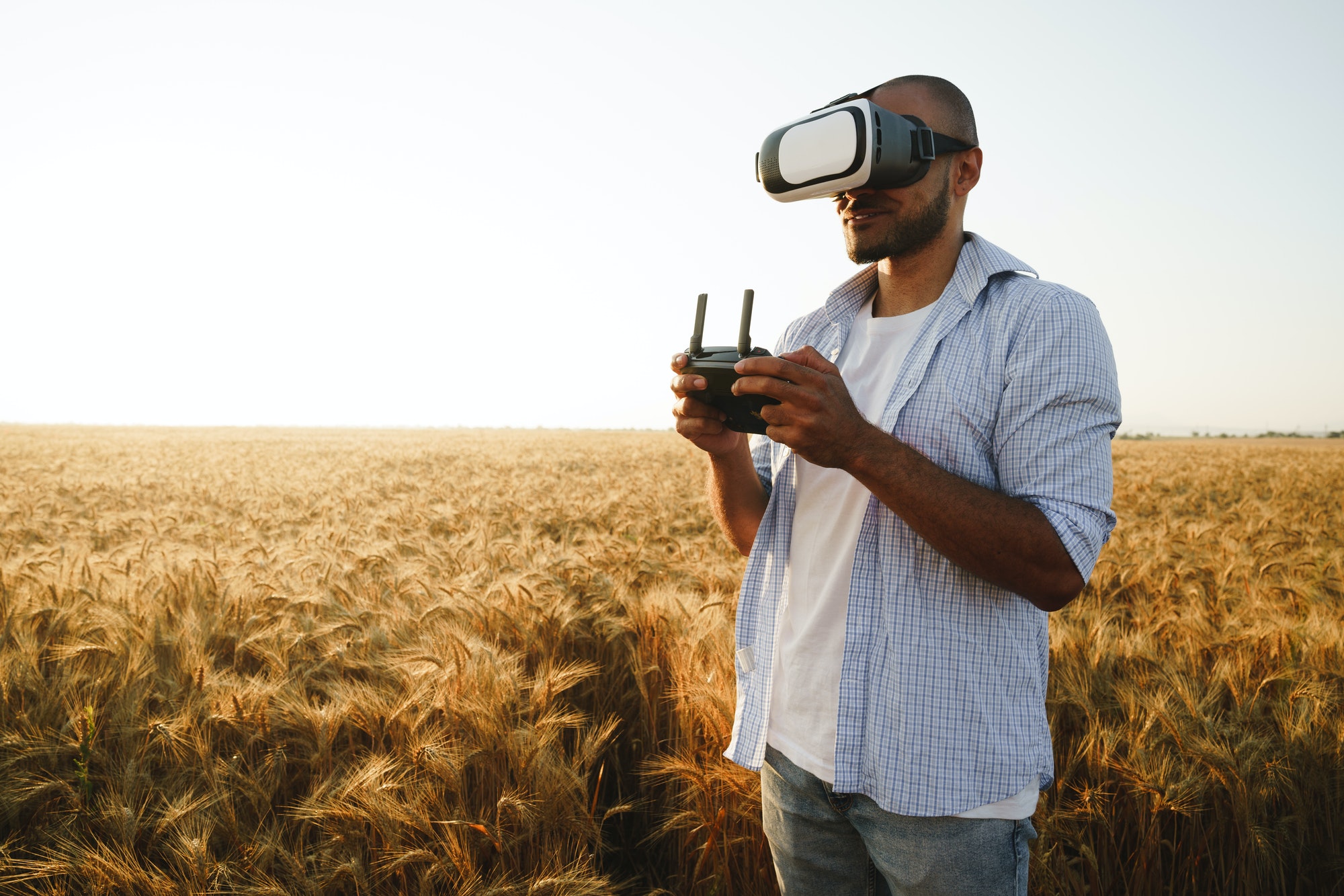 Young man standing in a wheat field at sunset in virtual reality glasses
